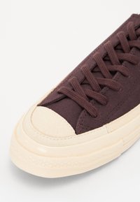 Hnědé plátěné tenisky s béžovou gumovou špičkou a podešví, s kulatými tkaničkami, texturovaným povrchem a prošíváním po stranách.