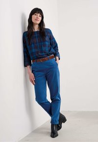 Sinine ruuduline särk nööpidega, pruun vöö, sinised corduroy püksid ja mustad pahkluu saapad. Mudel toetub lihtsale valgele seinale.