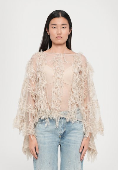 Châle beige transparent avec des détails en franges texturées. Coupe ample avec des manches larges, superposé sur un haut clair et associé à un jean délavé.