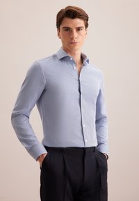 Camicia a maniche lunghe a scacchi blu e bianchi con colletto a punta, patta con bottoni e polsini, indossata con pantaloni blu navy, mani nelle tasche.