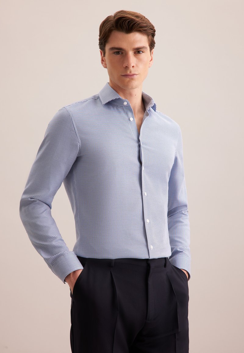 Camicia a maniche lunghe a scacchi blu e bianchi con colletto a punta, patta con bottoni e polsini, indossata con pantaloni blu navy, mani nelle tasche.