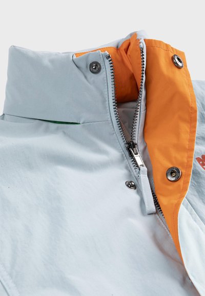 Col montant de veste bleu clair avec fermeture éclair argentée partiellement ouverte, doublure intérieure orange et boutons-pression argentés.