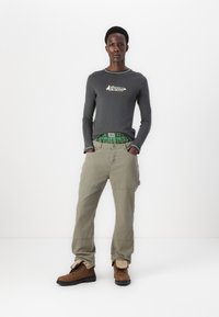 Γκρι μακρυμάνικο πουκάμισο με λογότυπο, πράσινη καρό ζώνη, ανοιχτό μπεζ cargo παντελόνι και καφέ μποτάκια με μαύρες σόλες, τοποθετημένα σε λευκό φόντο.