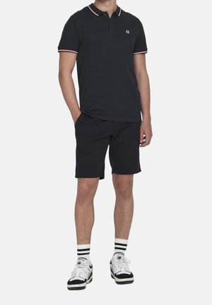 Schwarzes Polo-Shirt mit roten und weißen Akzenten, kombiniert mit schwarzen Shorts. Das Modell trägt weiße Sneaker mit schwarzen Akzenten und gestreifte Socken.
