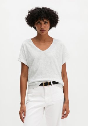 Kobieta o kręconych włosach ubrana w białą koszulkę z dekoltem w serek wpuszczoną w białe spodnie z czarnym paskiem, stojąca na jednolitym tle.