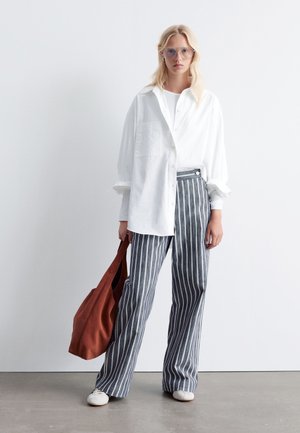 White button-up shirt, relaxed fit, long sleeves. Navy and white striped high-waisted trousers. Brown tote bag, white shoes. Neutral background.