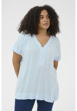 Femme aux cheveux bouclés portant un haut bleu clair à manches courtes et col en V, avec un jean bleu foncé, sur un fond uni.