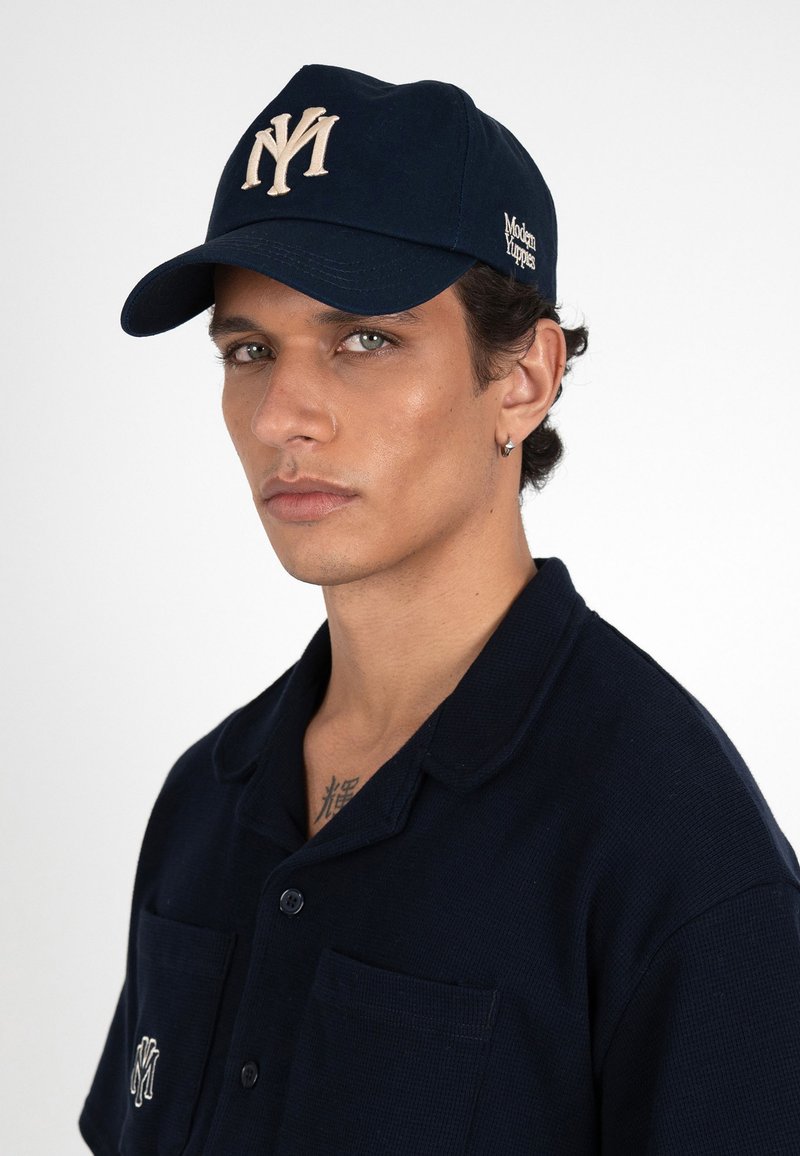 Jeune homme aux yeux clairs portant une casquette et une chemise bleu marine, toutes deux avec des logos blancs brodés, regardant légèrement sur le côté.