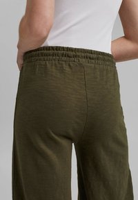 Pantaloni verde oliva con vestibilità rilassata, vita elastica e tessuto morbido e testurizzato. Un dettaglio di cucitura corre verticalmente lungo la schiena.