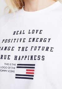 Felpa bianca con testo nero che presenta frasi e un logo. Il testo include "AMORE REALE", "ENERGIA POSITIVA" e un design a strisce colorate.