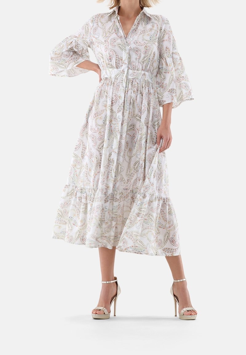 Femme porte une robe blanche à manches longues, longue jusqu'aux chevilles, avec un léger motif paisley et des sandales à talons beiges, debout, main sur la hanche.