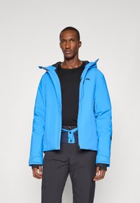 Casaco isolante azul claro com capuz, apresentando um logótipo preto, fecho com zíper e punhos ajustáveis. Usado sobre uma blusa e calças pretas.