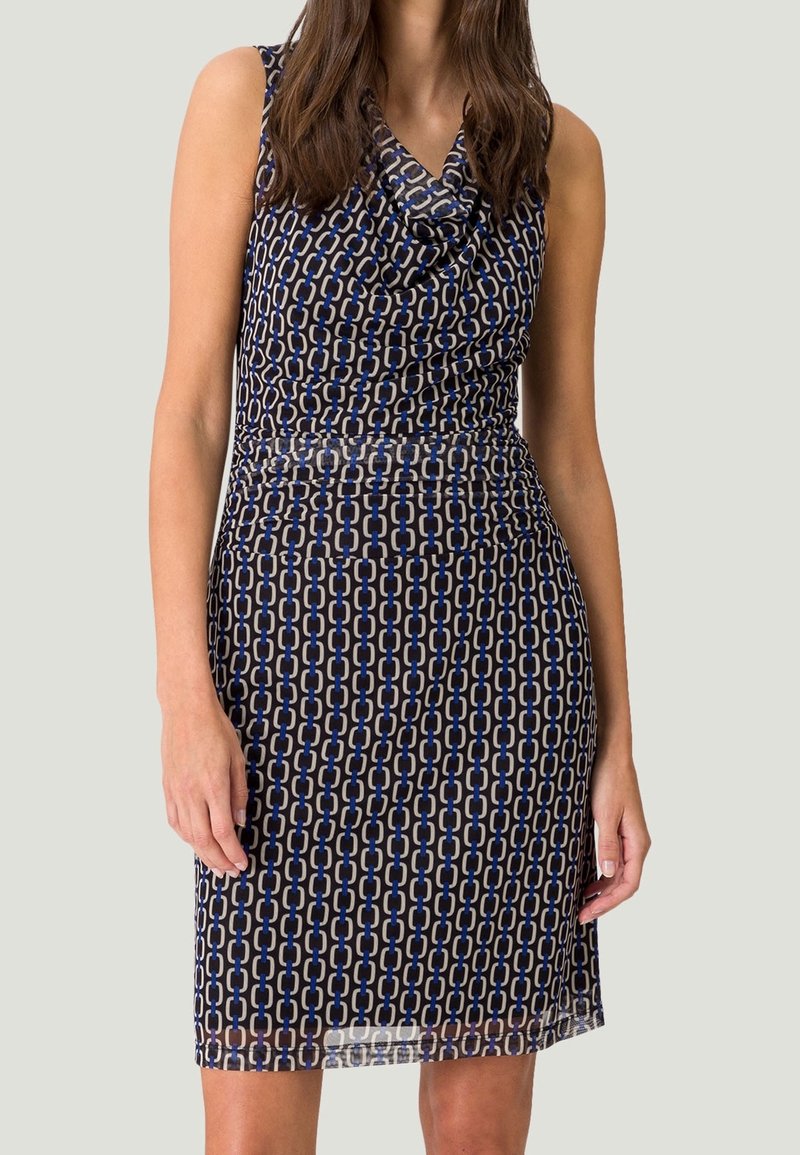 Femme portant une robe sans manches longueur genou avec un motif de chaînes liées noir, bleu et blanc et un col drapé.