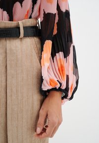 Långärmad, veckad blus med ett rosa och svart blommönster; har en elastisk manschett. Matchad med högmidjade, ribbade beige byxor.