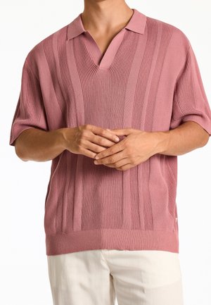 Mann trägt rosa kurzärmeliges Strick-Poloshirt mit vertikalen Rippenmustern und weiße Hose, Hände locker auf Taillenhöhe verschränkt.