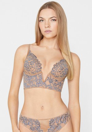 Femme blonde portant de la lingerie beige avec des motifs de dentelle florale bleue, debout devant un fond blanc uni.