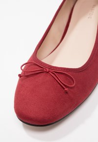 Ballerine en daim rouge à bout rond, avec un petit nœud assorti et une semelle noire contrastante. Intérieur lisse.