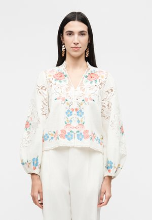 Kobieta nosząca białą bluzkę z różowym, niebieskim i żółtym kwiatowym haftem oraz koronkowymi detalami, w połączeniu z białymi spodniami i złotymi kolczykami.