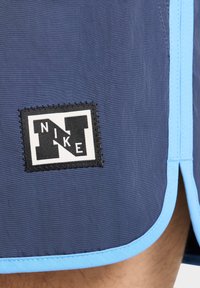 Mėlyni sportiniai šortai su juodu ir baltu siuvinėtų Nike logotipo pavidalu. Savybės: šviesiai mėlyni akcentai ir tekstūruotas audinio apdaila.