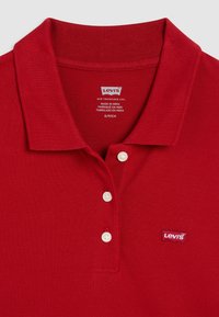 Červené polo tričko vyrobené z texturované bavlny s ribbeným límcem, obsahující tři bílé knoflíky a malé logo Levi's na hrudi.