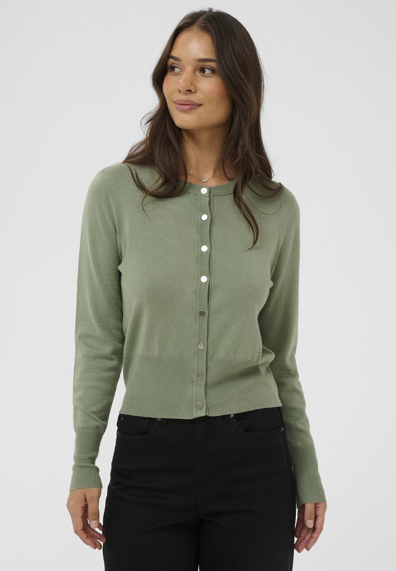 Naine, kes kannab salvei rohelise nööpidega kardigani ja musti pükse, vaatab küljele lõdvestunud hoiakus ja on taustal kerge heleda taustaga.