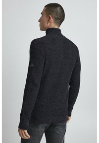Dunkelgrauer gestrickter Pullover mit hohem Kragen, strukturiertem Muster und langen Ärmeln, ausgestattet mit einem kleinen Logopatch am linken Arm.