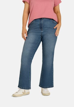 Person iført medium blå vidde-jeans, hvide sneakers og en lyserød kortærmet skjorte, stående med den ene hånd i lommen.