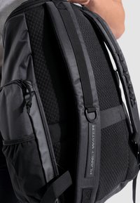 Zaino nero con tessuto strutturato, spallacci imbottiti e una tasca laterale. Marchio ben visibile sulla cinghia. Design minimalista.