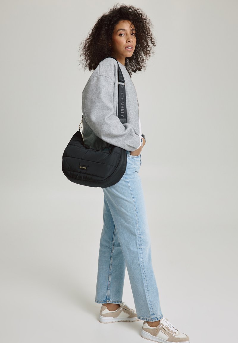 Schwarze gepolsterte Schultertasche mit einer geschwungenen Form, ausgestattet mit einem breiten Schultergurt und einem goldfarbenen Logodesign, getragen zu einem grauen Sweatshirt und hellen Jeans.