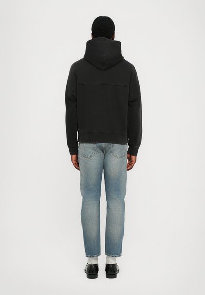 Schwarzer Kapuzenpullover mit lockerem Schnitt, aus Baumwollmaterial und mit einer strukturierten Oberfläche, kombiniert mit hellblauen schmal geschnittenen Jeans und schwarzen Schuhen.