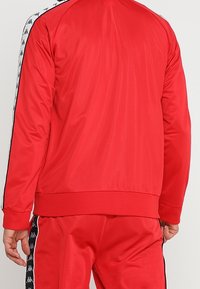 Rote Sportjacke aus glattem, leichtem Material. Verfügt über schwarze und weiße Streifenakzente entlang der Schultern und Ärmel.