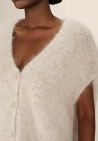 Gilet en tricot gris clair avec un col en V, des manches courtes et cinq boutons. Texture douce et duveteuse avec une coupe décontractée et des détails de couture subtils.