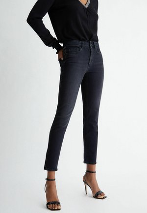 Femme portant un jean slim noir, un haut à manches longues noir et des sandales à talons hauts noires, se tenant avec une main dans la poche.
