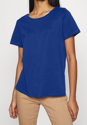 Kobieta ubrana w gładki, królewsko niebieski t-shirt z krótkim rękawem i beżowe spodnie, stojąca na tle jednolitego jasnego tła.