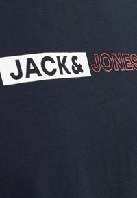 Marineblauwe katoenen t-shirt met een witte rechthoekige patch waarop "JACK&" in zwart en "JONES" in rood is afgebeeld, met een soepele textuur en een ronde halsontwerp.