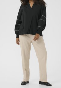 Blouse noire à encolure en V, manches ballon texturées avec coutures blanches, associée à un pantalon beige et des chaussures noires sans lacets.