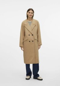Cappotto beige doppiopetto con grandi rever, due tasche frontali e bottoni neri, indossato sopra una maglietta a righe e abbinato a jeans a gamba larga.