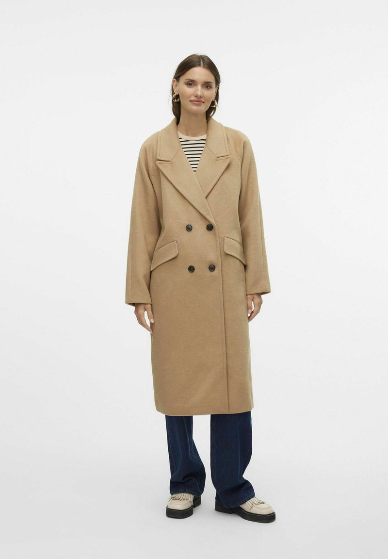 Cappotto beige doppiopetto con grandi rever, due tasche frontali e bottoni neri, indossato sopra una maglietta a righe e abbinato a jeans a gamba larga.