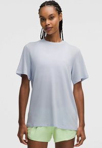 Vrouw met gevlochten haar, gekleed in een losse lichtgrijze T-shirt met korte mouwen en een lichtgroene korte broek, staand tegen een eenvoudige witte achtergrond.