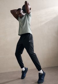 Schwarze Sporthosen und ein hellgrünes T-Shirt mit kurzen Ärmeln, das eine lockere Passform hat. Blaue Turnschuhe mit weißen Akzenten; Fitness-Studio.