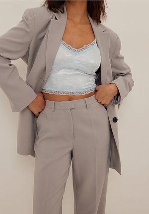 Frau trägt hellgrauen Anzug mit Blazer und Hose über einem blassblauen, mit Spitze verzierten, kurzen Top, Hände in die Hüften gestützt.