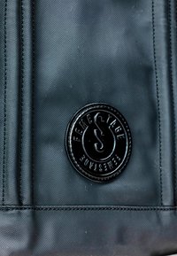 Czarna, teksturowana tkanina z okrągłą, błyszczącą naszywką logo na środku, z wytłoczonym tekstem i stylizowanym wzorem litery.