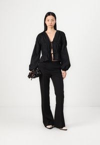 Fekete, rövid fazonú blúz mély V kivágással, ráncolt részletekkel és megkötős zárással. Fekete, kidolgozott nadrággal és mintás textil táskával párosítva.