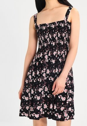 Frau trägt ärmelloses schwarzes Kleid mit rosa-weißem Blumenmuster und gerafftem Oberteil, steht vor hellem einfarbigem Hintergrund.