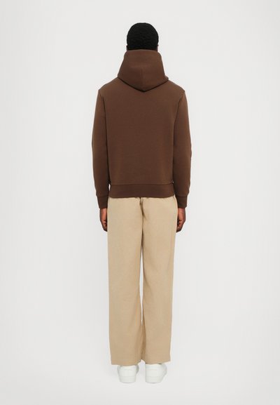 Sweatshirt capuz castanho com um corte descontraído, emparelhado com calças largas bege e ténis brancos, visto de costas.