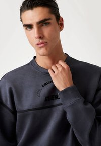 Donkere grijze sweatshirt gemaakt van zacht materiaal met een ronde hals, zwarte accenten en bedrukte logo's. Model poseert met een casual houding.