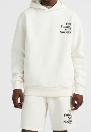 Personne portant un sweat à capuche blanc et un short avec un texte noir indiquant "The Future Surf Society" sur la poitrine et la cuisse, sur un fond uni.