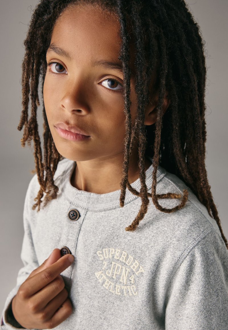 Grijze gebreide trui met een ronde hals, voorzien van twee donkere knopen en een crème-kleurig geborduurd logo op de borst. Gestructureerde stof en een comfortabele pasvorm.