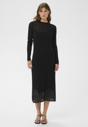 Schwarzes geripptes Midi-Kleid mit langen Ärmeln und transparenter Textur, mit hohem Kragen und figurbetontem Schnitt. Akzentuiert mit minimalistischem Schuhwerk.