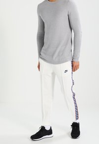 Grå långärmad stickad tröja kombinerad med vita sweatpants som har en blå och röd mönstrad sidremsa. Svarta träningsskor fulländar outfiten.